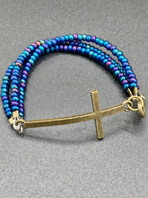 Load image into Gallery viewer, For Positivity and Faith, Shimmery Blue Glass Beaded Bracelet With Curved Horizontal Brass Cross. This bracelet has a large brass cross that is slightly curved to be comfortably worm around your wrist. I created three glass beaded strands that have a wide variety of blue, purple, green, and gold tones. Measurements: 7 1/2&quot; in length, brass cross measures 2&quot; in length
