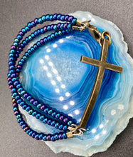 Load image into Gallery viewer, For Positivity and Faith, Shimmery Blue Glass Beaded Bracelet With Curved Horizontal Brass Cross. This bracelet has a large brass cross that is slightly curved to be comfortably worm around your wrist. I created three glass beaded strands that have a wide variety of blue, purple, green, and gold tones. Measurements: 7 1/2&quot; in length, brass cross measures 2&quot; in length
