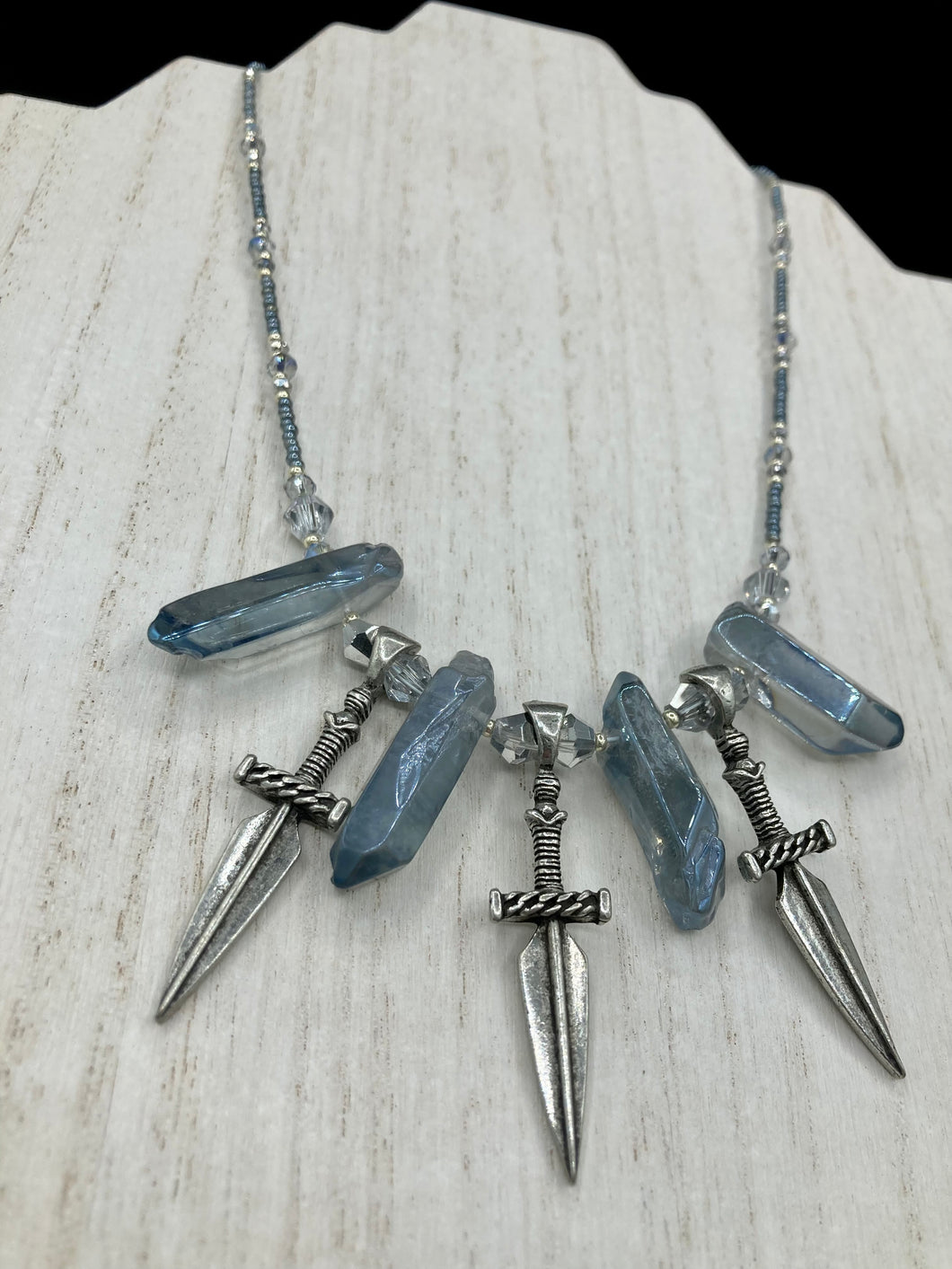 This elegant and dark necklace features shimmering light silver and gray glass beads of varying sizes and shapes for texture and dimension. The necklace is finished with blue luster crystal stone nuggets and nickel-free dagger pendants, creating a beautiful draping effect. Measurements:   Necklace is 17”adjustable up to 19