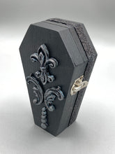 Load image into Gallery viewer, This vintage-inspired coffin ring box will make a spooky yet cute addition to any jewelry collection. The sleek black box has eye-catching black and gray embellishments, while the interior is beautifully decorated with a vivid turquoise blue fabric and a highly detailed black and silver cross with dark red roses. Function and style meet with this unique and stylish piece.
