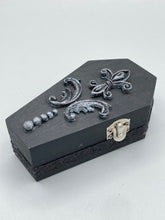 Load image into Gallery viewer, This vintage-inspired coffin ring box will make a spooky yet cute addition to any jewelry collection. The sleek black box has eye-catching black and gray embellishments, while the interior is beautifully decorated with a vivid turquoise blue fabric and a highly detailed black and silver cross with dark red roses. Function and style meet with this unique and stylish piece.
